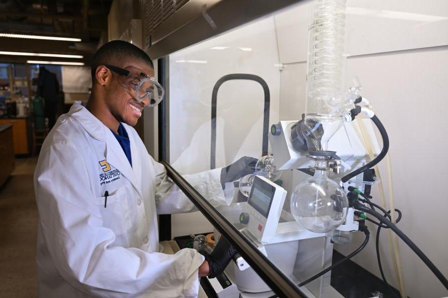 男联合技术大学化学系学生，戴着护目镜，穿着实验服，站在实验设备旁边